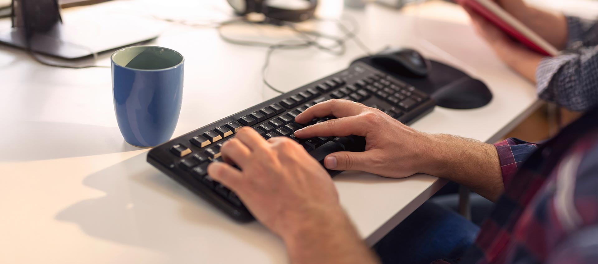 Hands type on a black keyboard on a white desk, with a blue mug nearby. The background shows blurred office items and a person holding a red book.