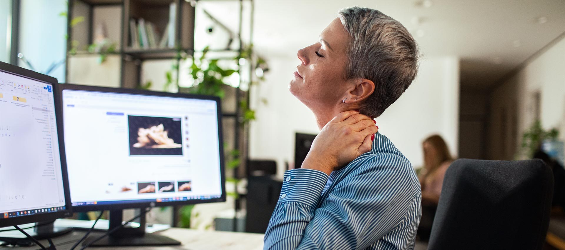 A person rubs their neck, appearing to feel discomfort, while sitting at a workspace with two computer monitors. Bookshelves and plants are visible in the background.