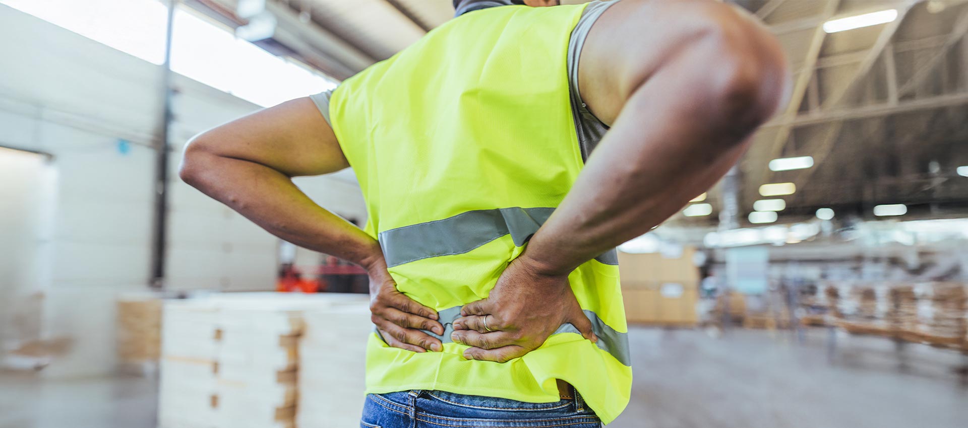 A person in a fluorescent safety vest stands with hands on their lower back in a warehouse, suggesting discomfort or pain.