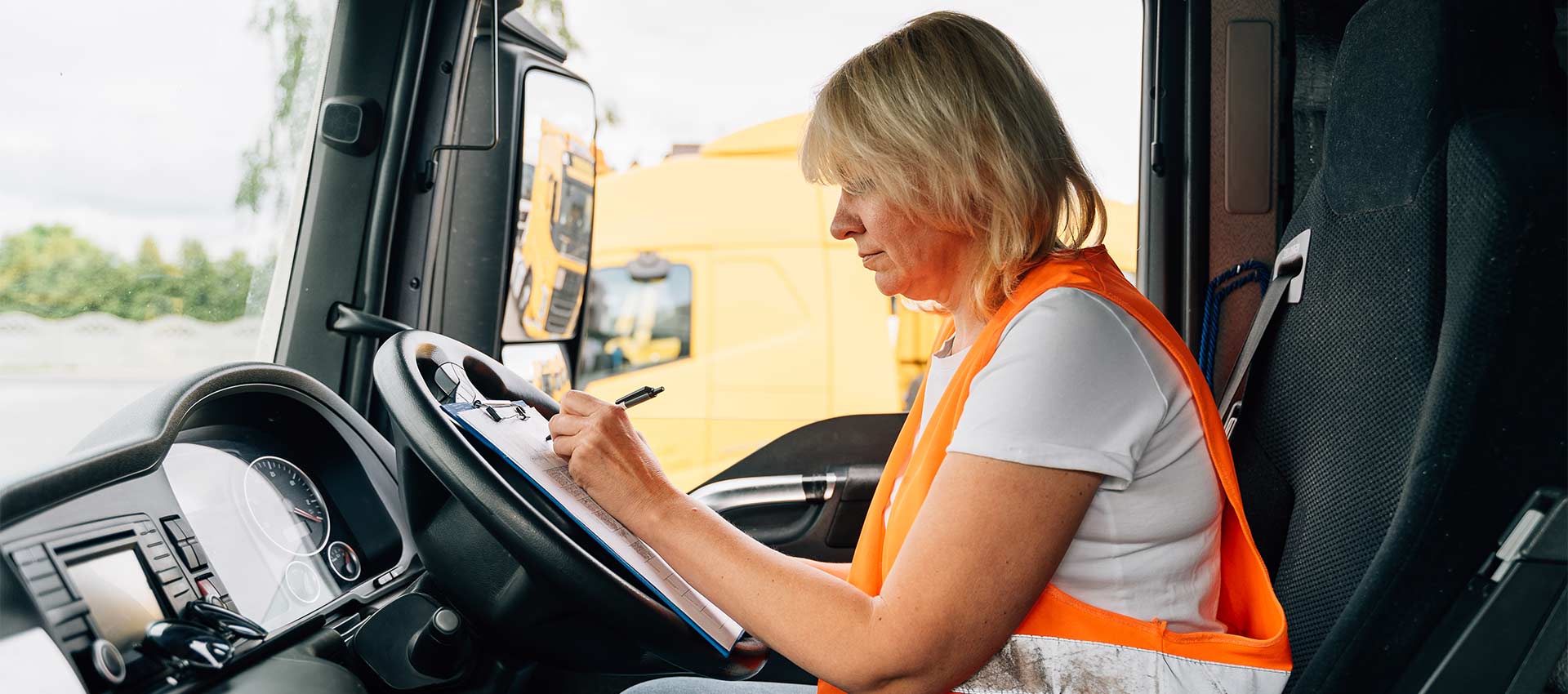 A person in an orange safety vest writes on a clipboard inside a parked truck cab, with a yellow vehicle visible through the window.