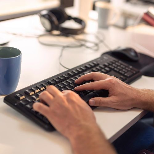 Hands type on a black keyboard on a white desk, with a blue mug nearby. The background shows blurred office items and a person holding a red book.