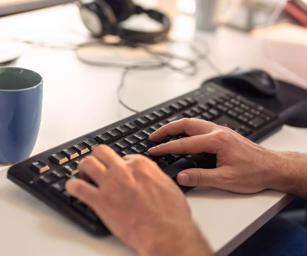 Hands type on a black keyboard on a white desk, with a blue mug nearby. The background shows blurred office items and a person holding a red book.