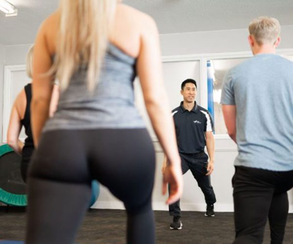 A fitness instructor guides a group of people during a workout session in a gym. Participants stand on blue mats, facing the instructor, with exercise equipment visible in the background.