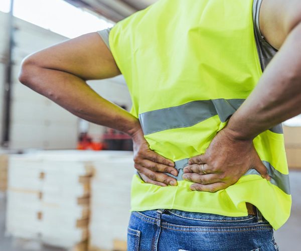 A person in a high-visibility vest holds their lower back in apparent discomfort, standing in a warehouse environment with stacked boxes in the background.
