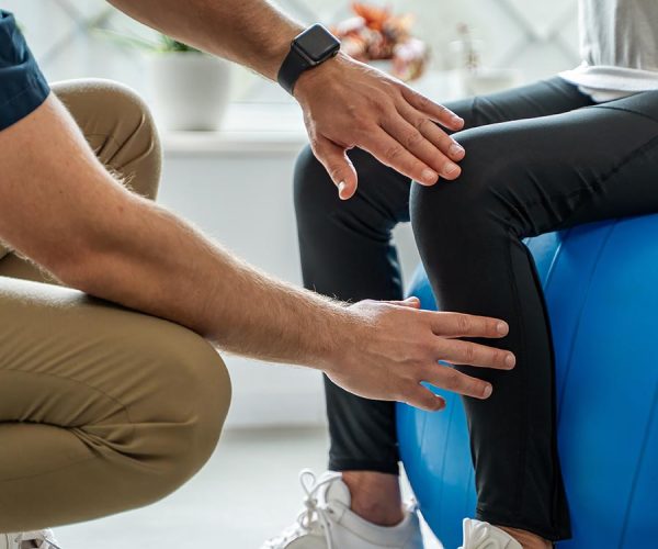 A healthcare professional assesses a seated person's knee, with hands positioned near it. The person sits on a blue exercise ball, and indoor plants are visible in the background.