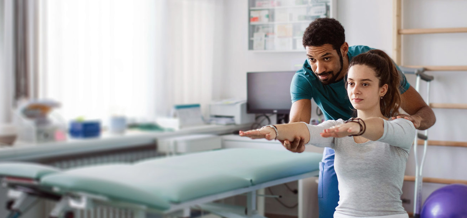 A physical therapist assists a seated woman with stretching exercises, extending her arms forward. They are in a bright, well-equipped therapy room with medical equipment and wall charts in the background.