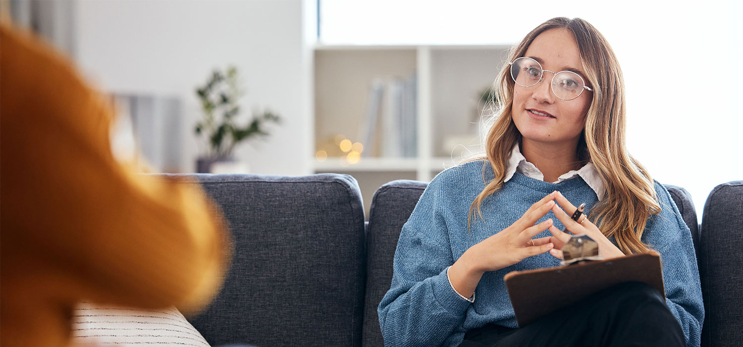 A person with glasses holds a clipboard and gestures thoughtfully while seated on a sofa in a well-lit room with a plant in the background.