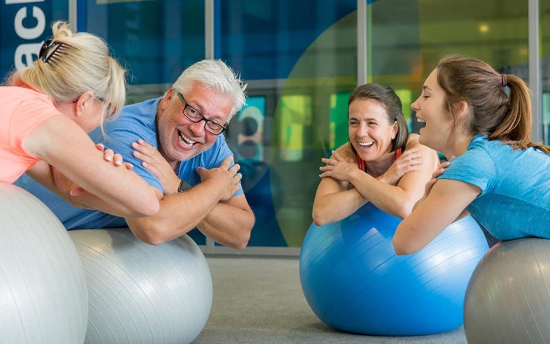 Four people lean on exercise balls, smiling and engaging in conversation in a bright gym setting, creating a lively and supportive atmosphere for physical fitness.