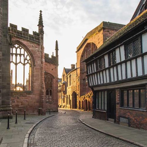 A cobblestone street curves between historical buildings with Gothic and Tudor architectural styles, under a softly lit sky, conveying a serene, old-world charm.