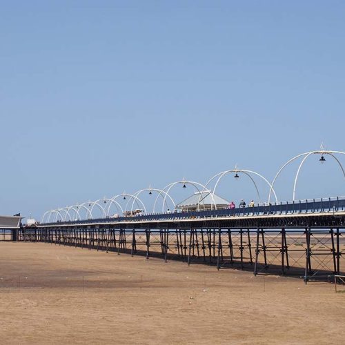 A long, elevated pier extends into the distance over a sandy beach, with arches and streetlights adorning its path. The sky is clear and blue.