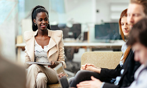 A healthcare professional holds a clipboard and smiles while speaking with two seated individuals in a modern office setting featuring a laptop and a window in the background.