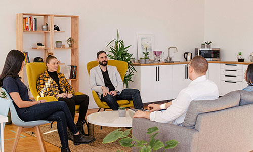A group of people sit around a small table in an office setting, engaged in conversation, with a potted plant and shelves in the background.