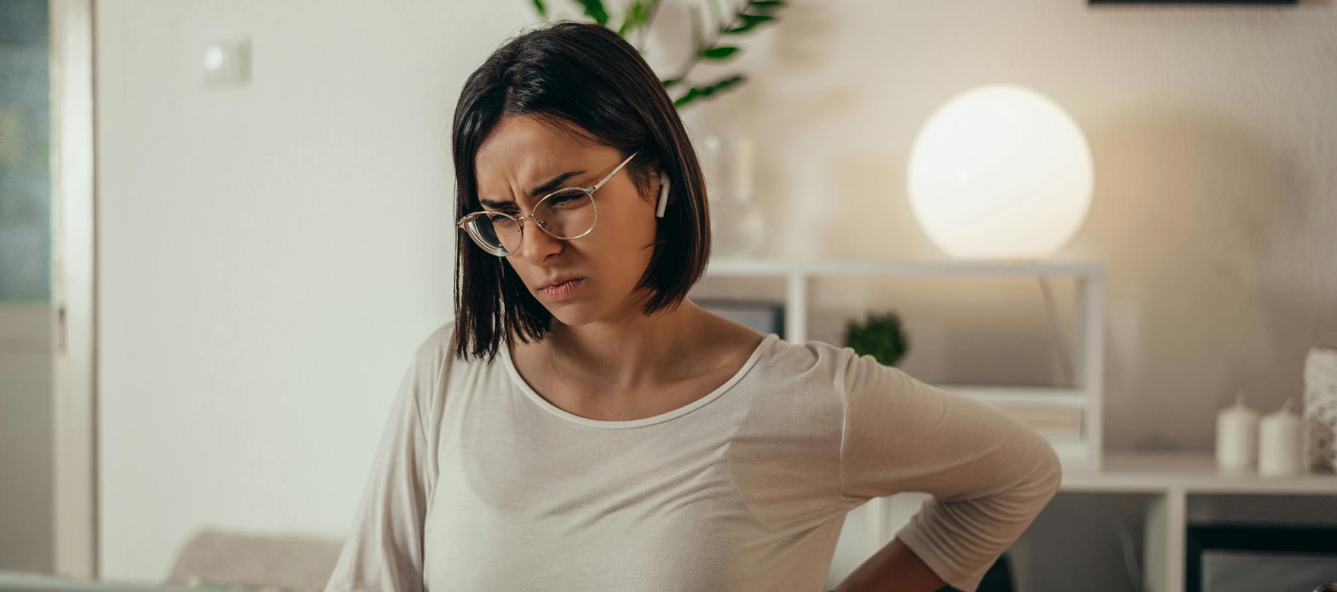A woman sitting at a disk in warmly lit room, holds her hand against her back.