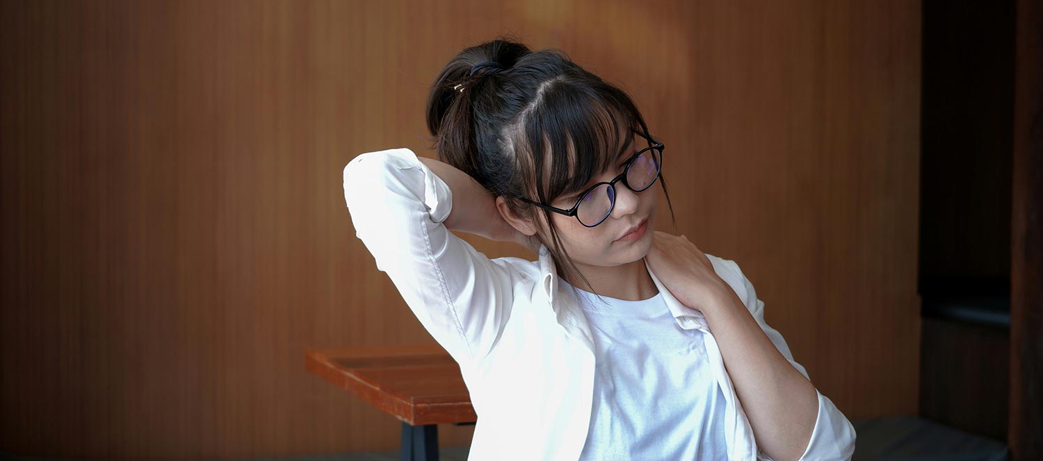 A woman wearing a white shirt, black glasses with her hair tied up in a pony tail and a full fringe, stretches her neck and massages her shoulder while sit at a desk.