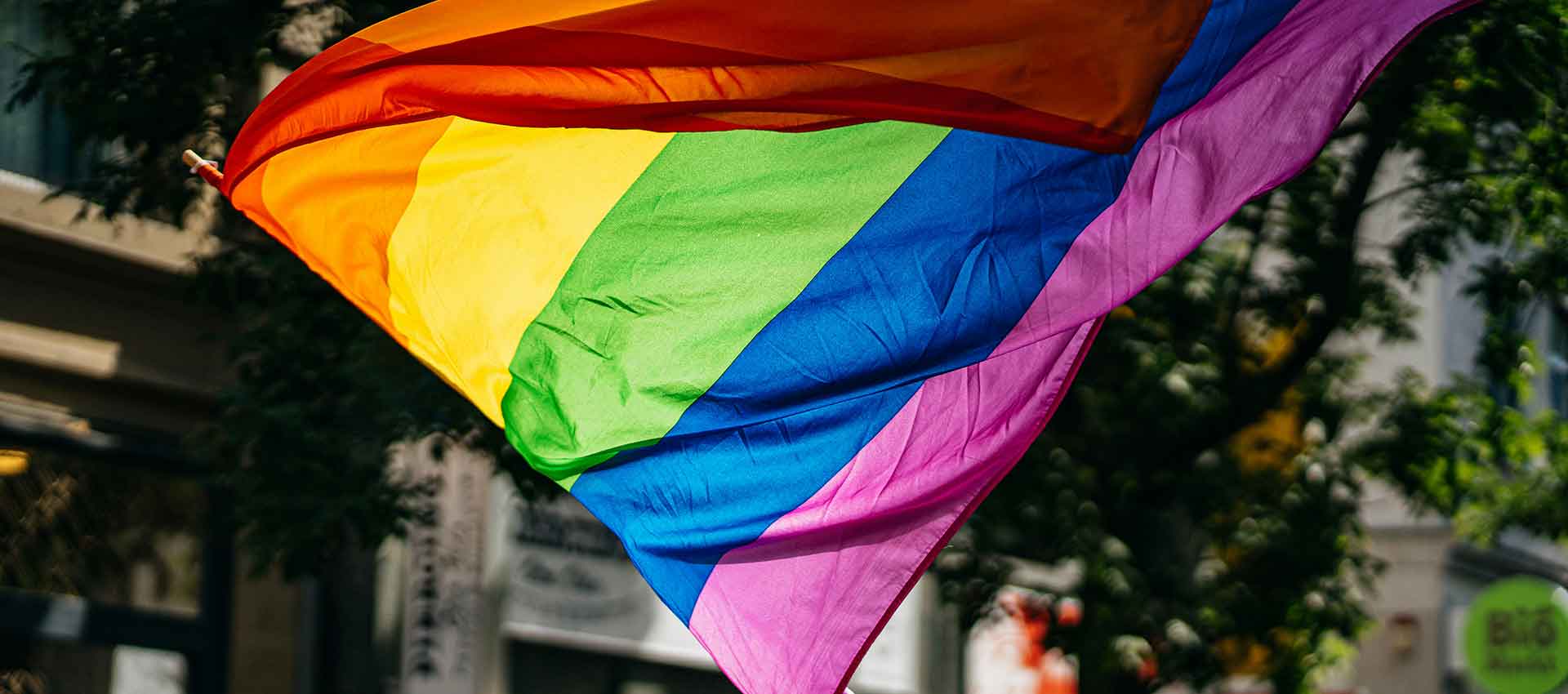 A pride flag made up of red, orange, yelllow, green, blue and purple stripes blows in the wind of a busy street.