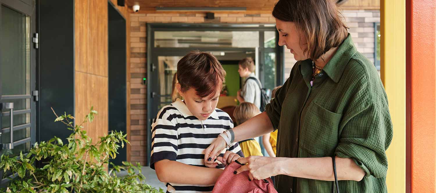 A mother helps her son put his thing inside a back pack as she drops him off at school.