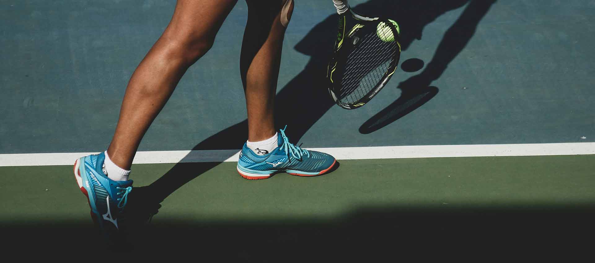 A close up of a woman with tanned legs stood on a tennis court, wearing tennis shoes and holding a tennis racket.