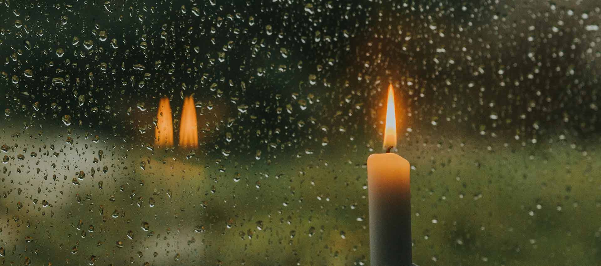 A close up of a lit flame on a tall thin candle that sits next to a window covered in rain drops.