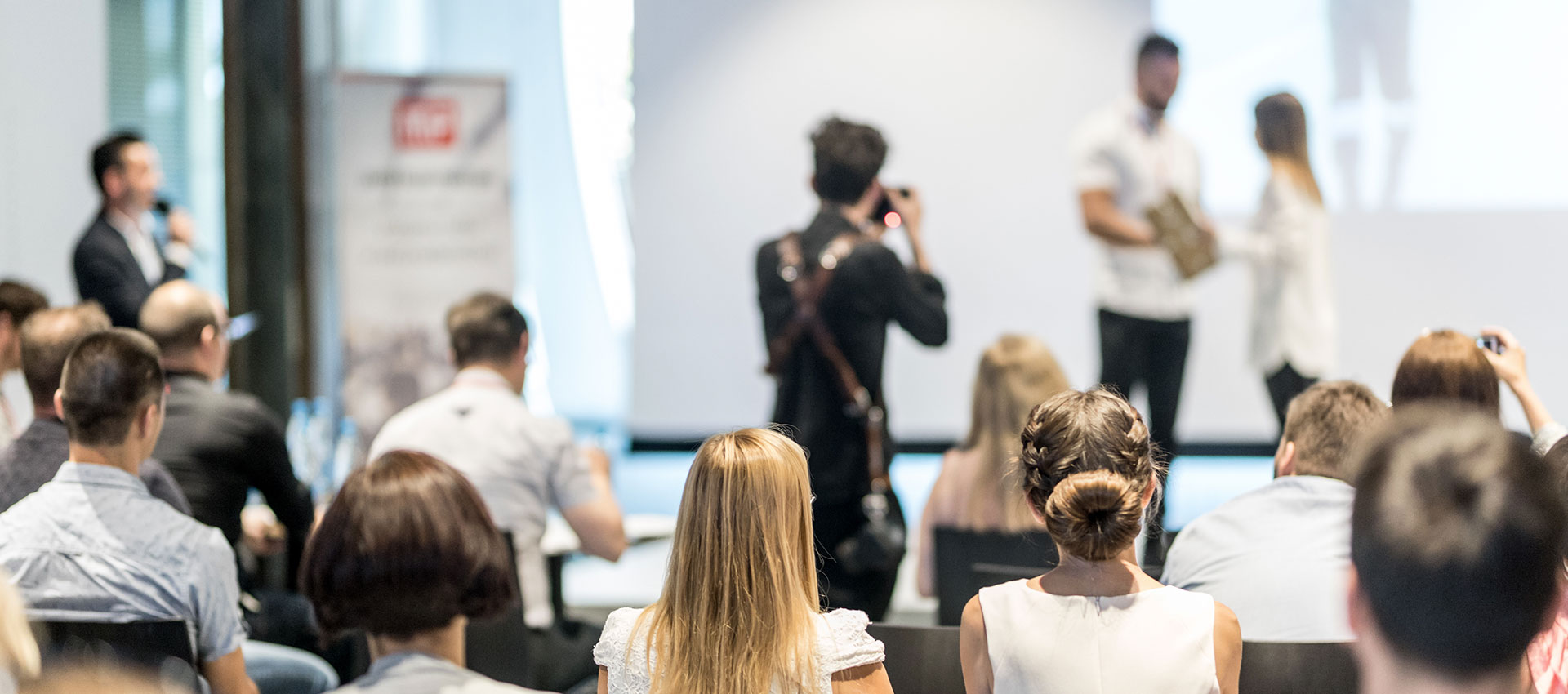 Audience members seated in a conference room watch a presentation, with two individuals standing and speaking in front of a bright screen. A banner is visible on the side.