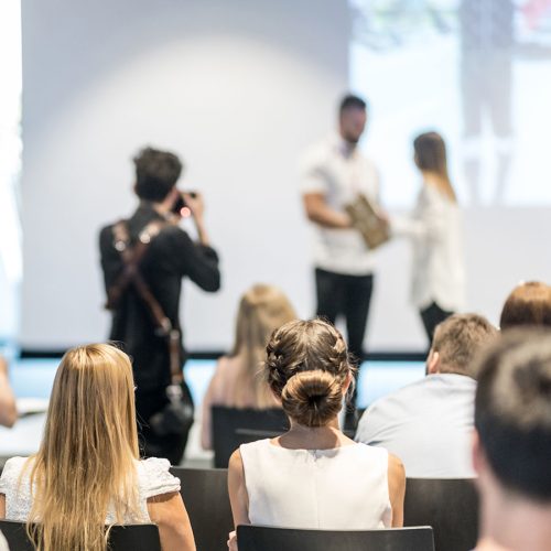 A person presents an award to another on stage while an audience watches. A photographer captures the moment in a bright conference room with a large screen in the background.