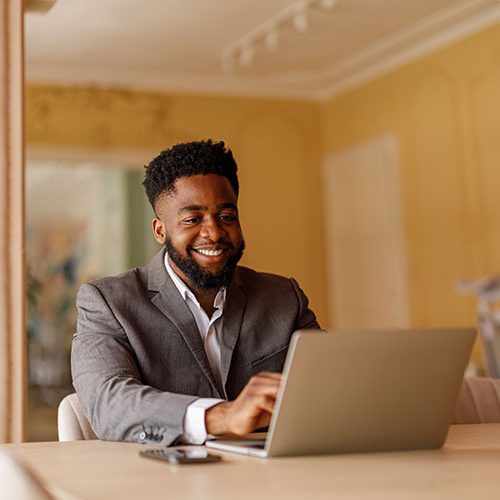 A man in a suit is typing on a laptop in a warmly lit office with beige walls and track lighting.