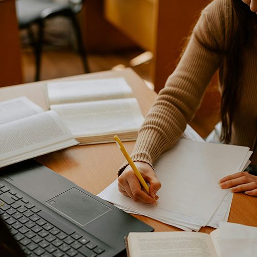 A person is writing notes with a yellow pencil on paper. Open books and a laptop are on the wooden desk in a well-lit room. No text is visible.