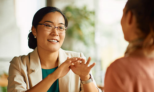 A person gestures expressively with hands while speaking to another person in a brightly lit room, suggesting a friendly and engaging conversation.