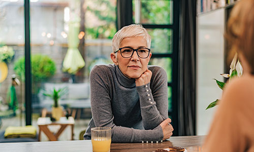 An individual with short gray hair and glasses sits at a table, resting their chin on their hand, with a glass of orange juice nearby, in a bright, modern room.