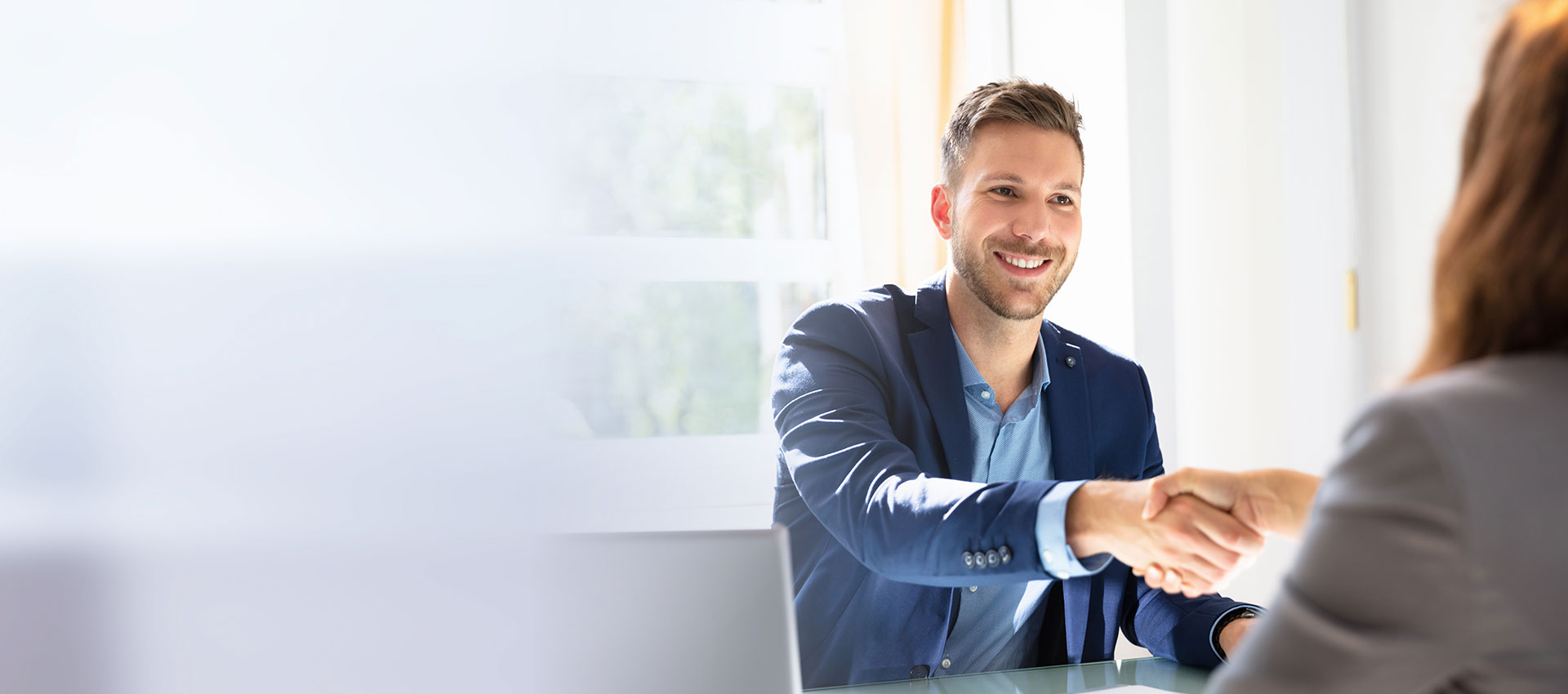A man in a blue suit is smiling and shaking hands with a woman in a bright office setting, suggesting a professional agreement or meeting.
