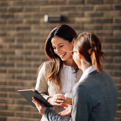 Two people stand outside against a brick wall. One holds a tablet and a cup, smiling while showing the screen to the other, who listens attentively.