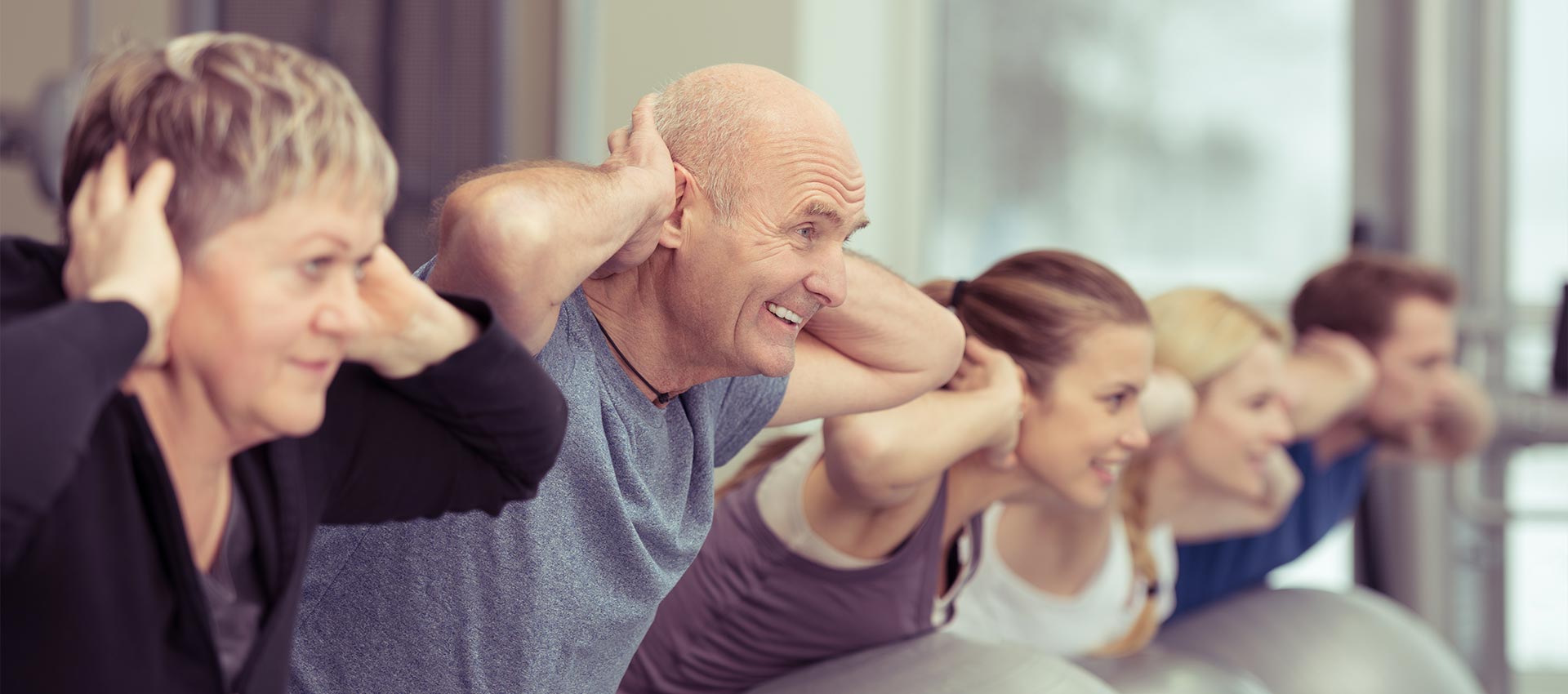 People exercise by leaning forward with hands behind their heads, balancing on large fitness balls, in a bright gym setting.