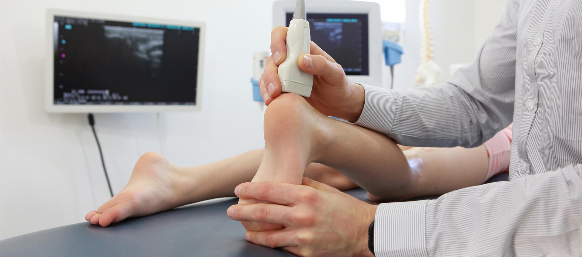 A healthcare professional conducts an ultrasound scan on a patient's foot in a medical setting, with ultrasound images displayed on monitors in the background.
