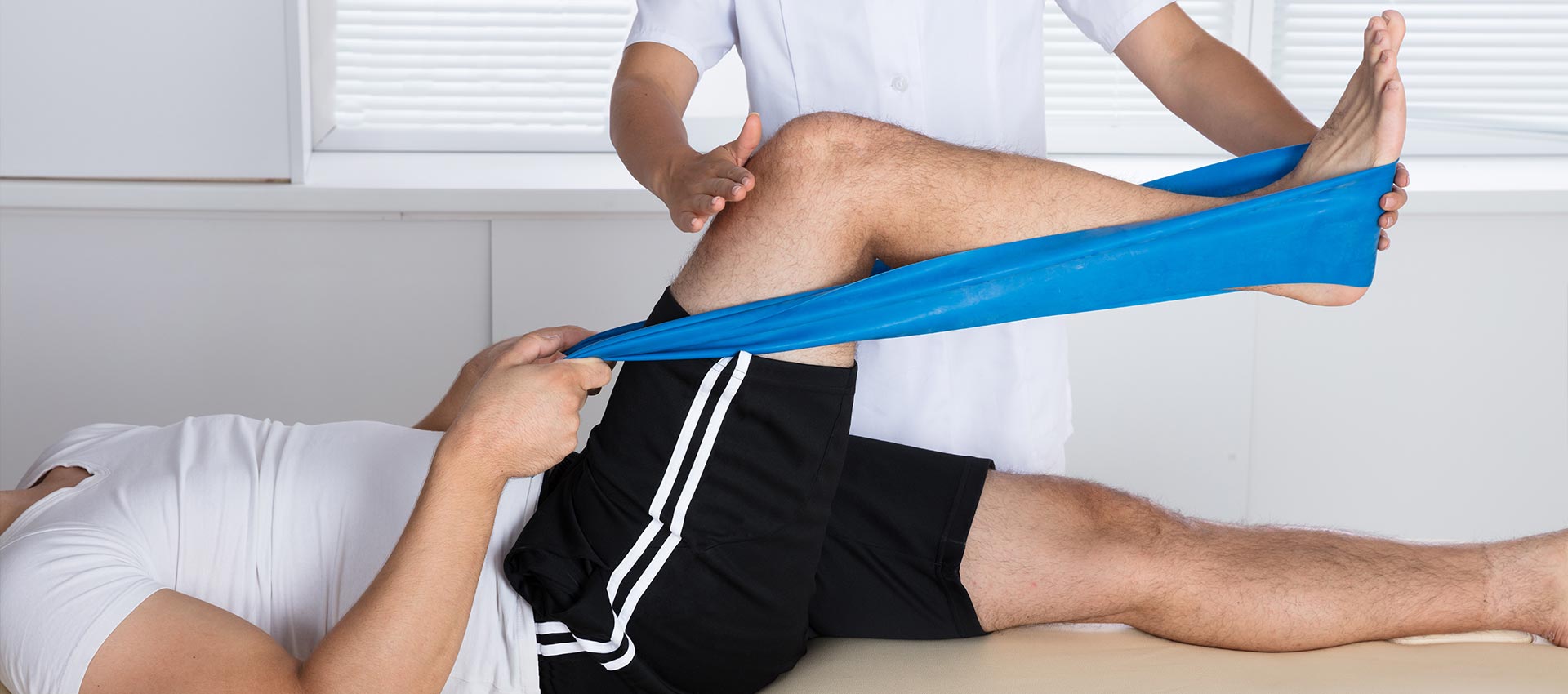 An older man exercises with a green resistance band, assisted by a younger person in a brightly lit room, suggesting a therapy or rehabilitation session.