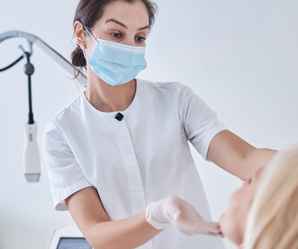 A healthcare professional wearing a face mask and gloves examines a patient in a clinical setting with medical equipment nearby.