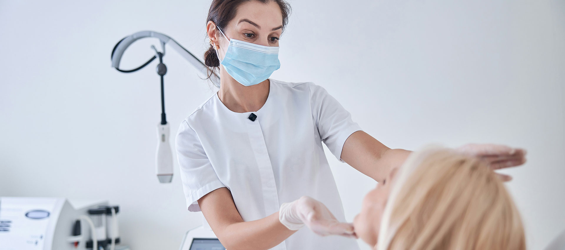 A healthcare professional wearing a face mask and gloves examines a patient in a clinical setting with medical equipment nearby.