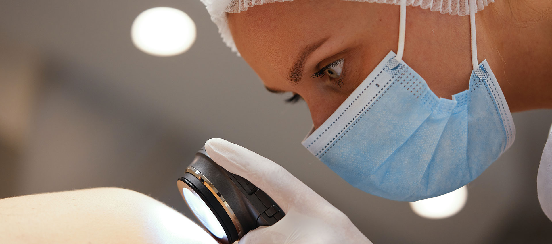 A healthcare professional closely examines a patient's skin using a dermatoscope. The person wears a surgical mask and gloves, with overhead lights softly illuminating the clinical environment.