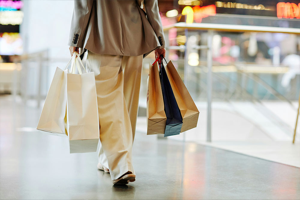 A person carrying several shopping bags walks through a mall with shiny flooring and blurred bright signs in the background.