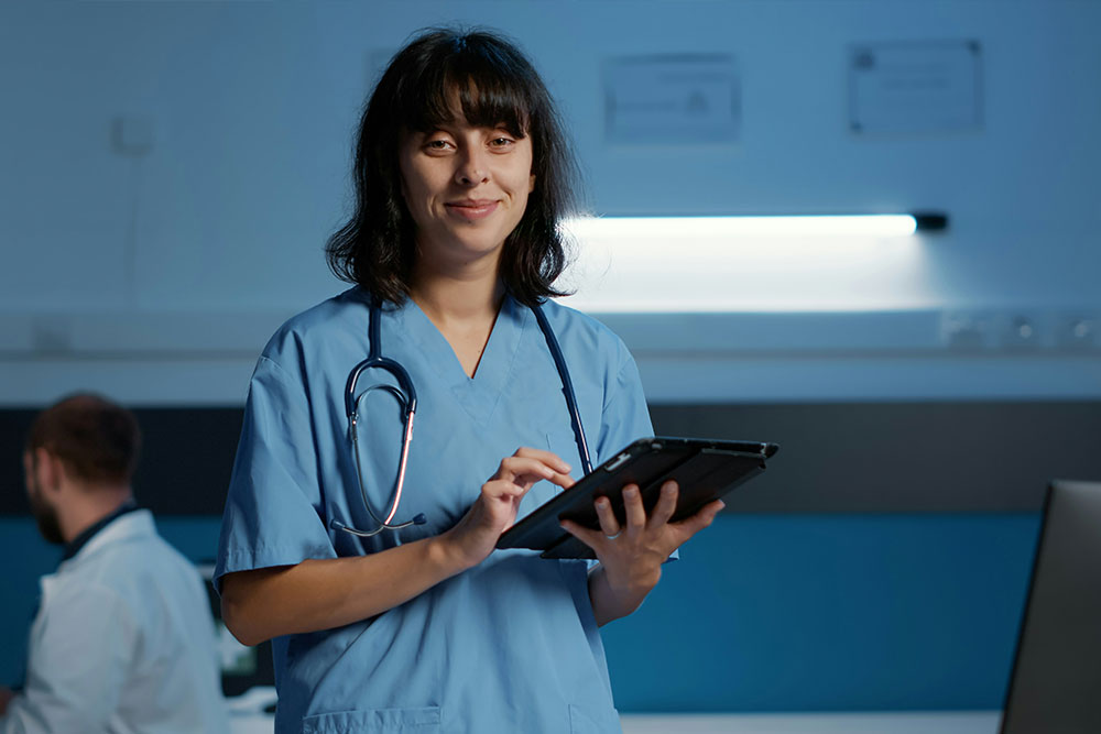 A healthcare professional in blue scrubs uses a tablet, with a stethoscope around their neck. They stand in a well-lit, modern healthcare setting, with another professional in the background.