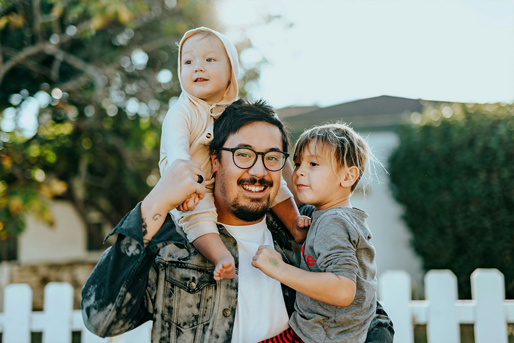 A person is holding two children, one on each arm, while standing in front of a white picket fence with greenery in the background, smiling in the sunlight.