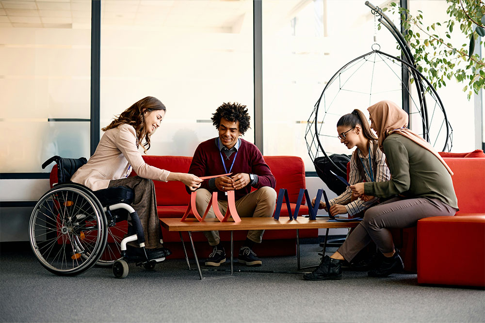 Four people sit around a low table in an office lounge, engaging in a collaborative activity with a decorative object. A wheelchair and hanging chair are visible in the bright room.
