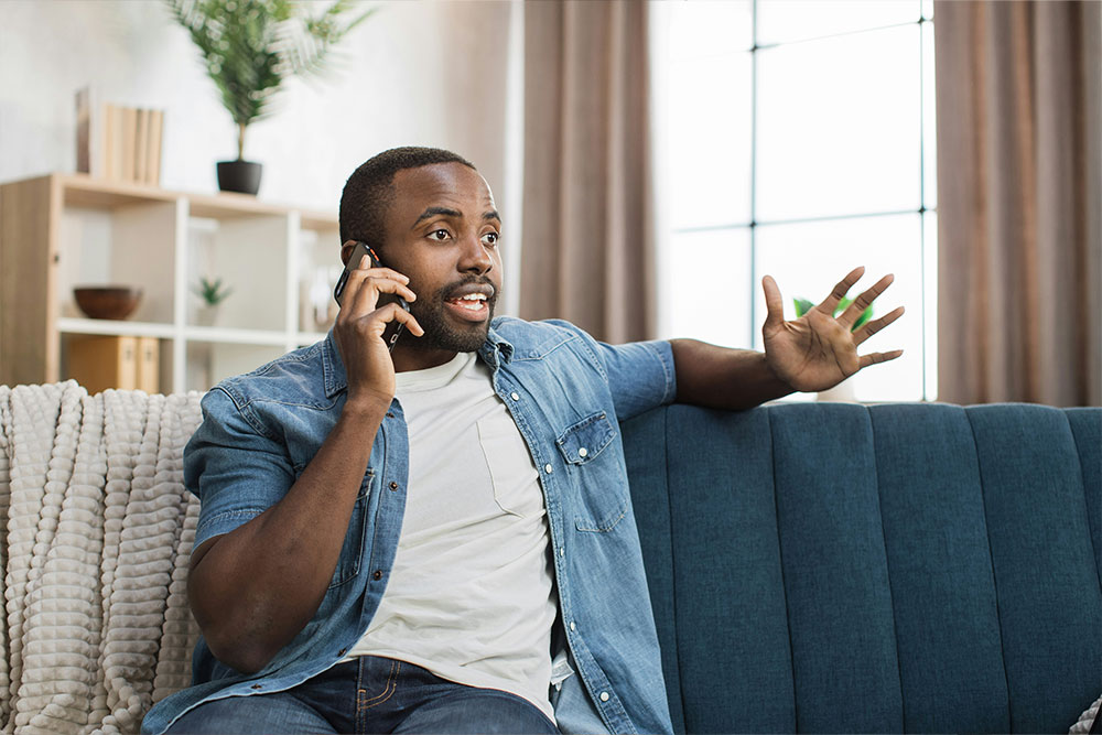 A man sits on a blue couch, talking on a smartphone and gesturing with his hand. Behind him, a shelf and a large window suggest a home setting.