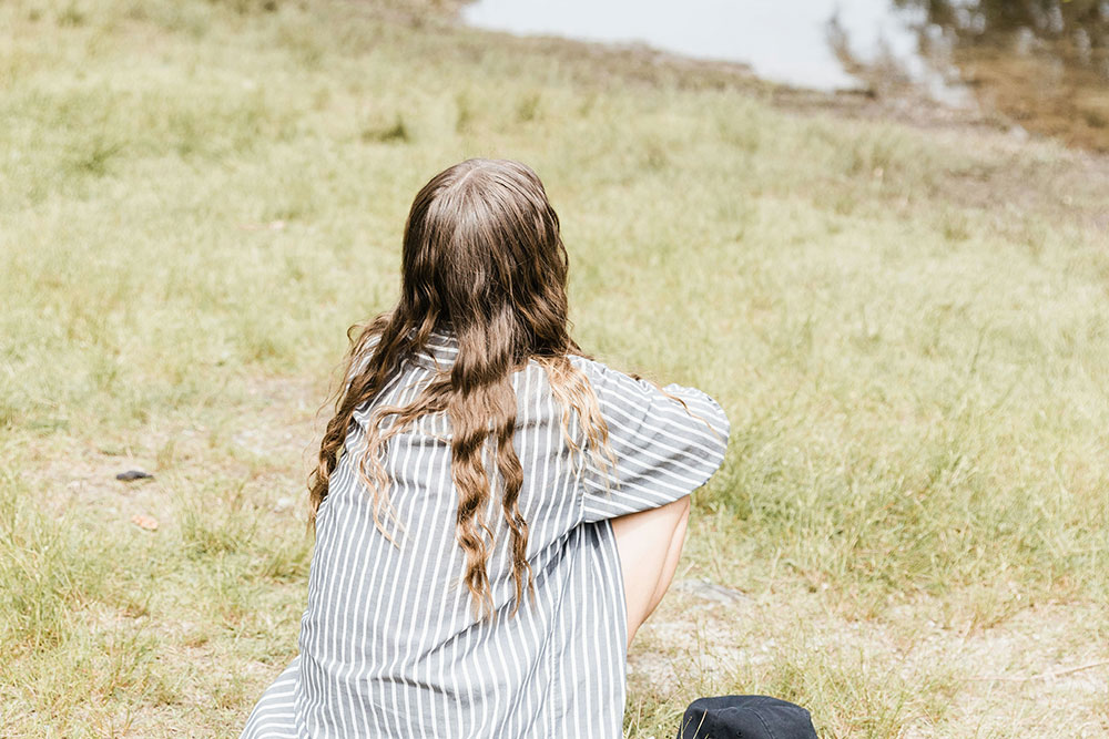 A person with long hair sits cross-legged facing away, wearing a striped shirt, on a grassy field near water.