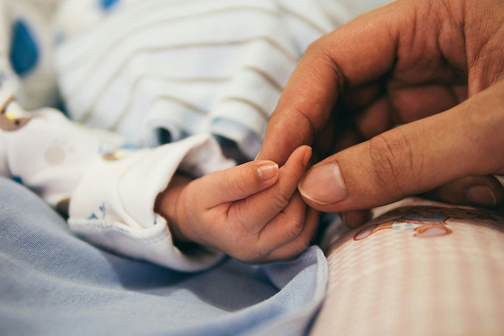Adult hand gently holding a baby's hand, both resting on blue and pink clothing. The baby wears a white, patterned outfit with stripes visible in the background.