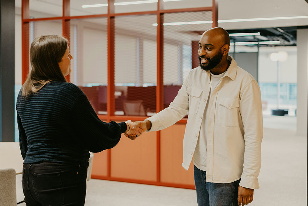 Two people are shaking hands in a modern office environment with red partition panels and bright lighting. They appear to be greeting each other warmly.