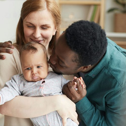 A woman and a man lovingly embrace and kiss a baby. The setting is a cozy home interior with soft lighting and plants in the background.