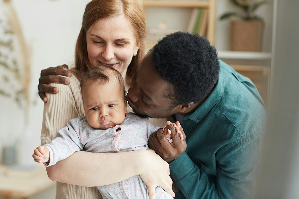 A woman and a man lovingly embrace and kiss a baby. The setting is a cozy home interior with soft lighting and plants in the background.