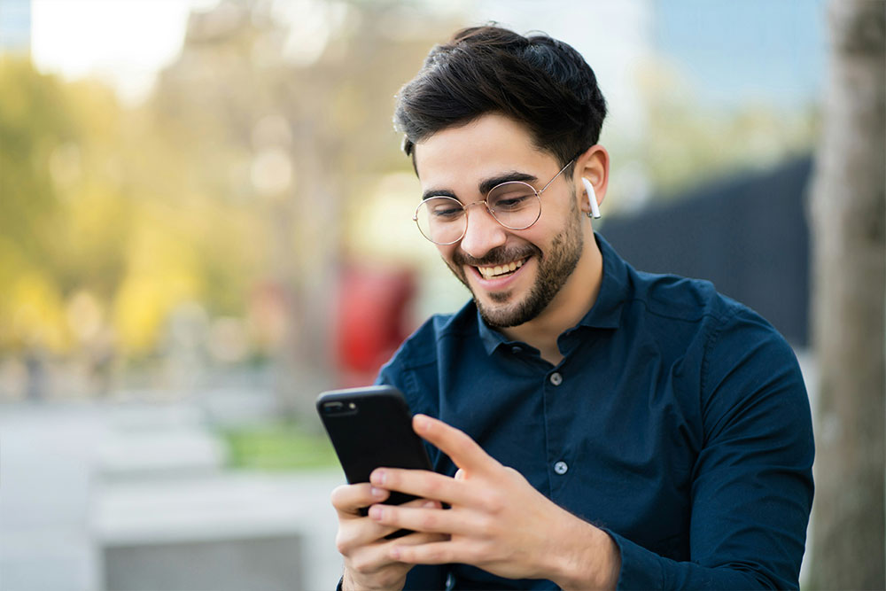 Man smiling while looking at a smartphone, wearing wireless earbuds and glasses, seated outdoors with blurred trees and buildings in the background.