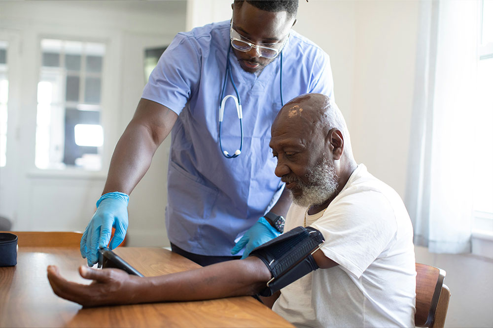 Healthcare professional in blue scrubs checks blood pressure of seated older man in a white shirt. They are in a bright, softly lit room with windows in the background.