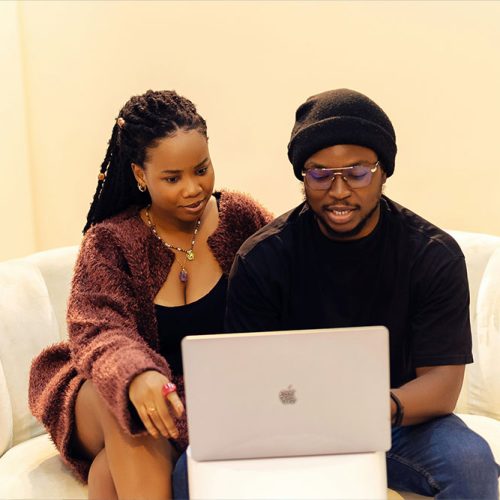 Two people sit together on a white sofa, focusing on a laptop screen. A potted plant is visible in the background, enhancing the warm, inviting atmosphere.