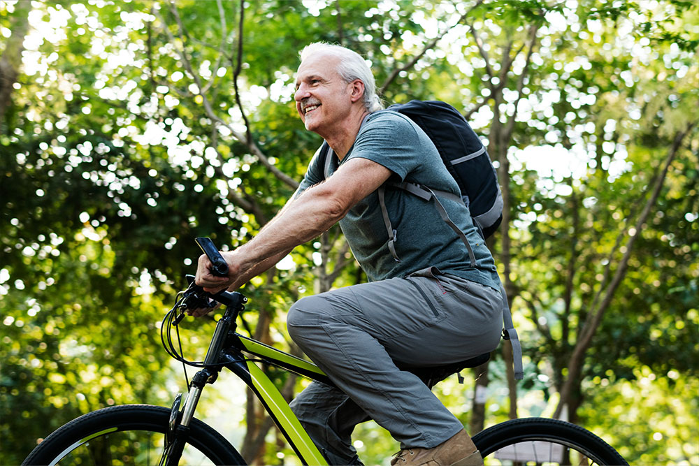 A man rides a bicycle through a sunlit forest path, wearing a backpack and casual clothing. He smiles, enjoying the outdoor activity amid lush green trees.